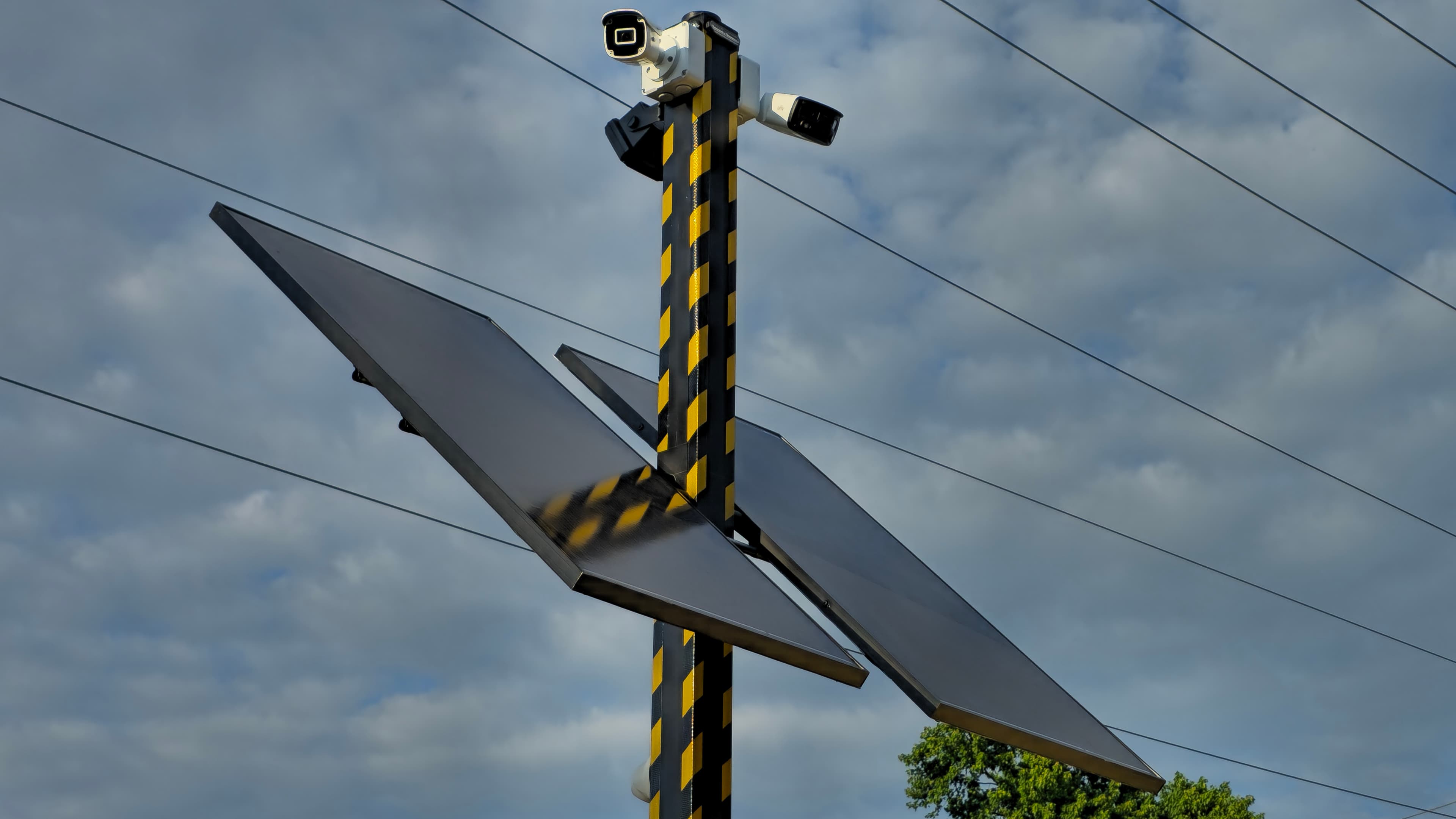 Solar panels and security cameras on a yellow and black striped pole against cloudy skies.